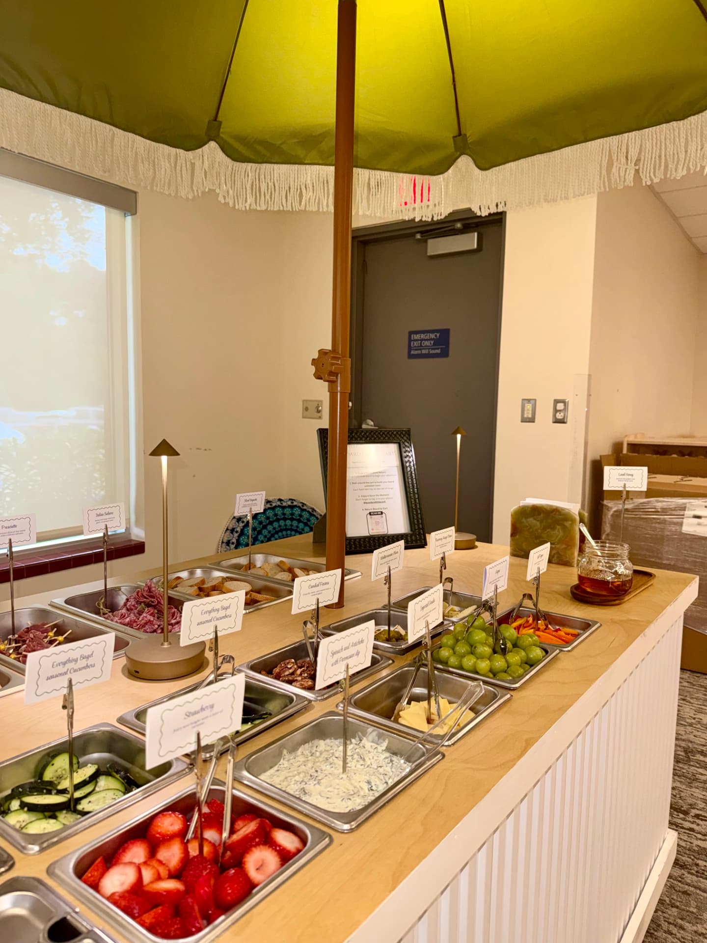 The charcuterie cart under a chartreuse fringed umbrella with labeled trays at a corporate event