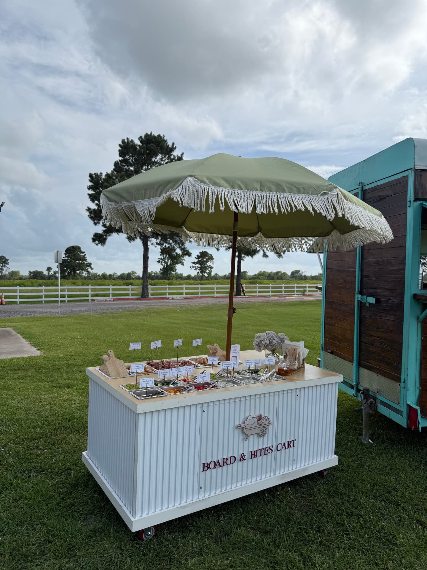 The Board & Bites mobile charcuterie cart outdoors under a green fringed umbrella